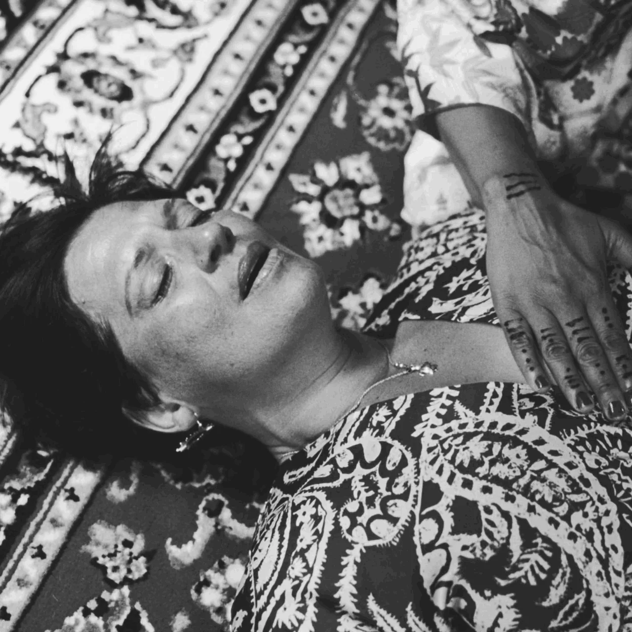 Anissa Lalahoum allongée sur un tapis à motifs, les yeux clos, une main tatouée au henné près du visage.