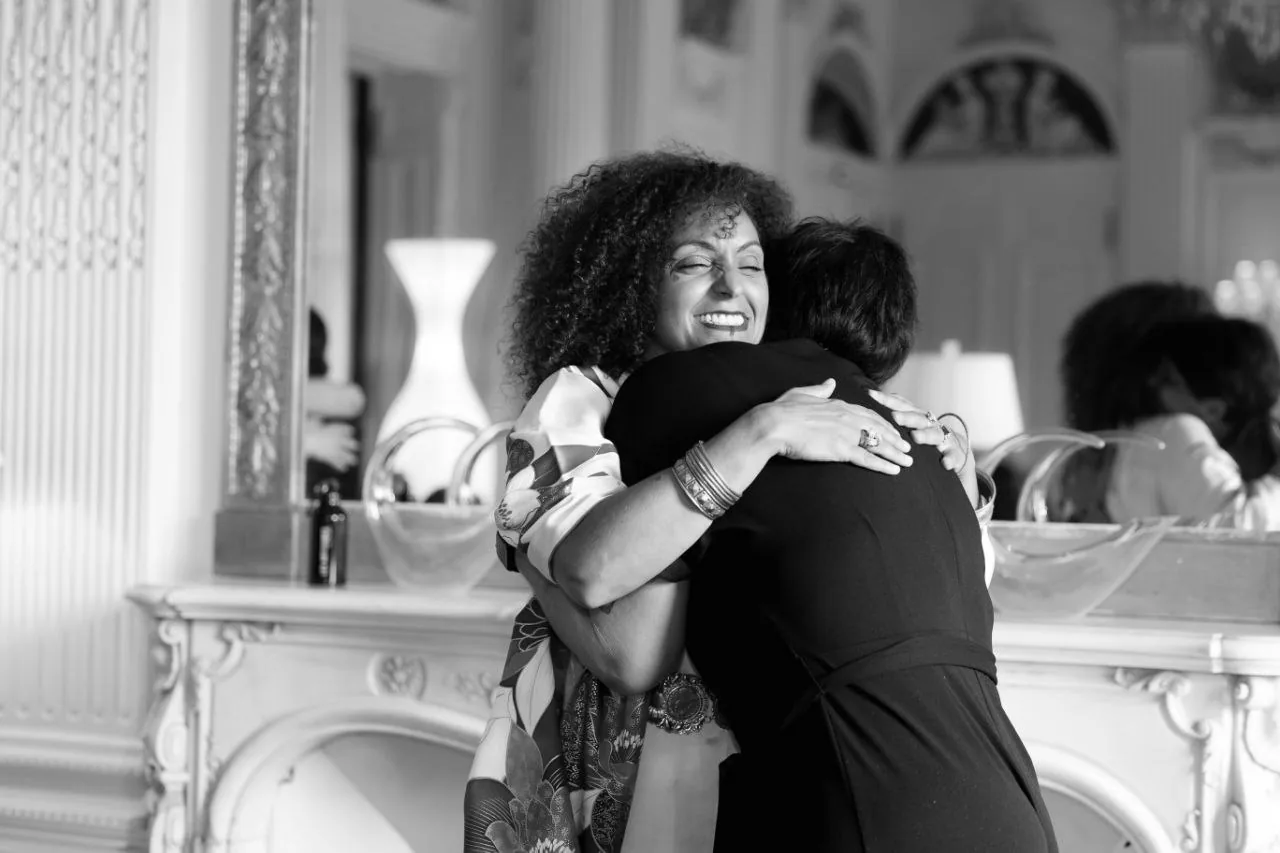 Anissa Lalahoum, souriante, enlace une femme vue de dos, dans un salon aux murs clairs.