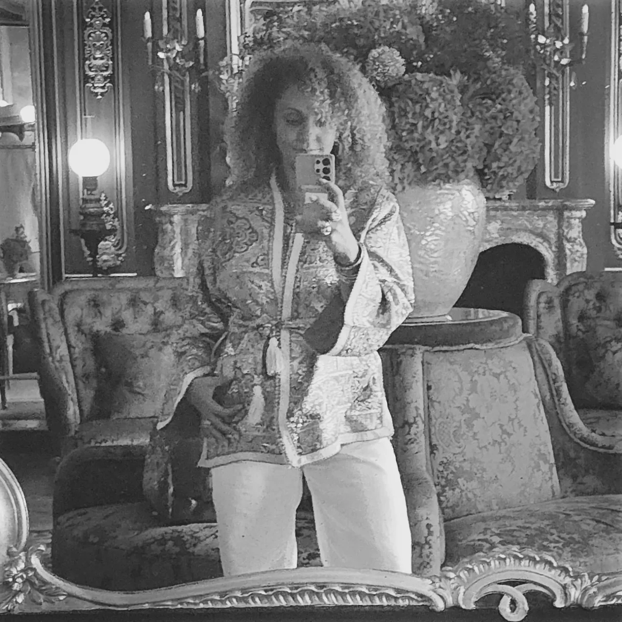 Anissa Lalahoum, en veste brodée, se photographie dans le miroir d’un salon aux lampes dorées.