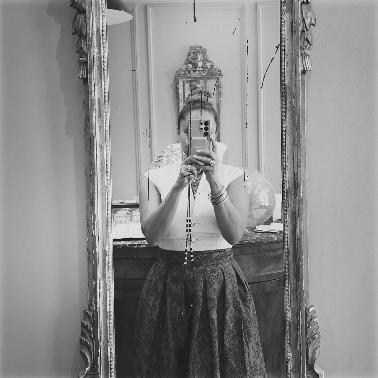 Anissa Lalahoum debout devant un grand miroir au cadre doré, le téléphone devant le visage, en jupe longue.