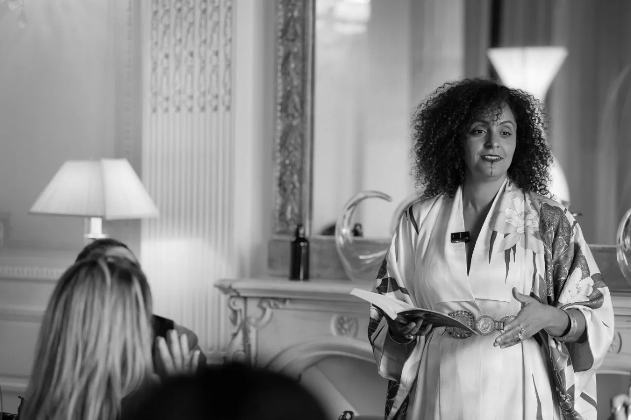 Anissa Lalahoum debout dans un salon aux moulures, en kimono, parlant à un groupe assis devant elle.