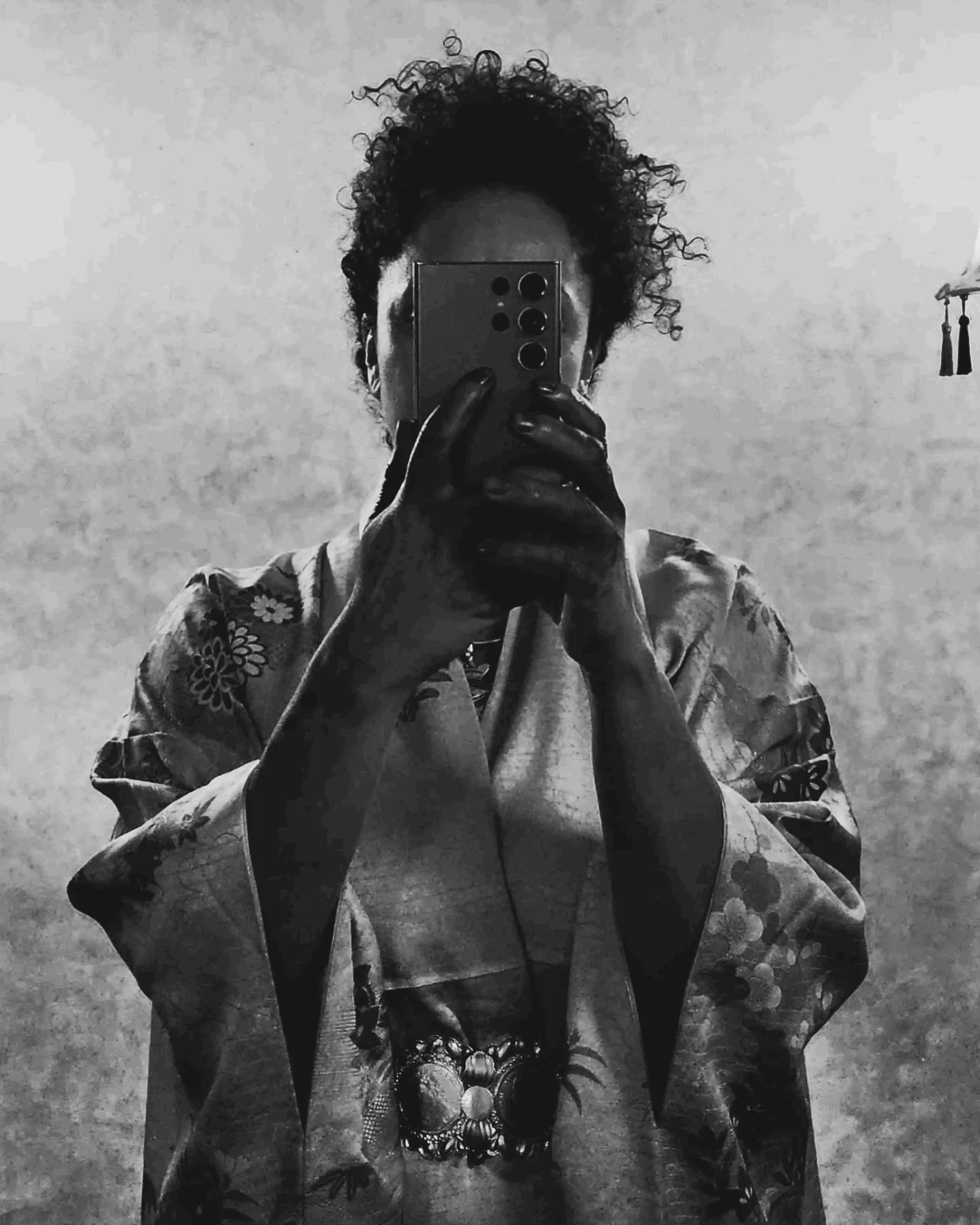 Anissa Lalahoum en kimono brodé, un téléphone tenu à deux mains devant le visage, en noir et blanc.