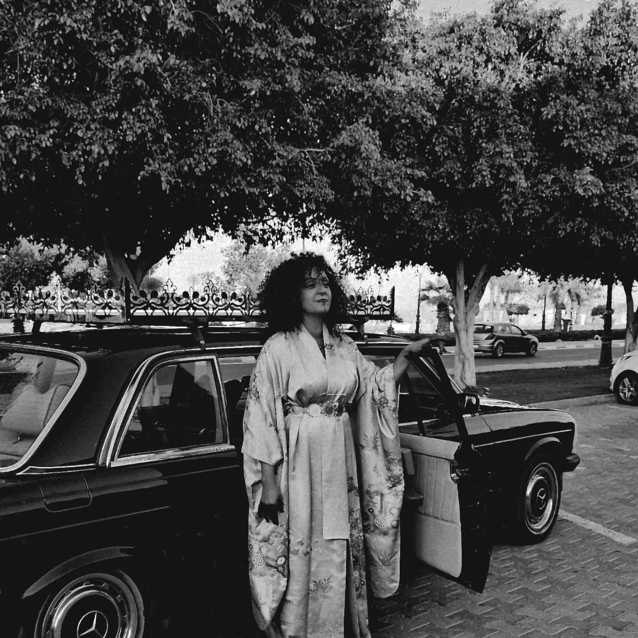 Anissa Lalahoum debout en kimono clair devant une voiture ancienne, sous les arbres, en noir et blanc.